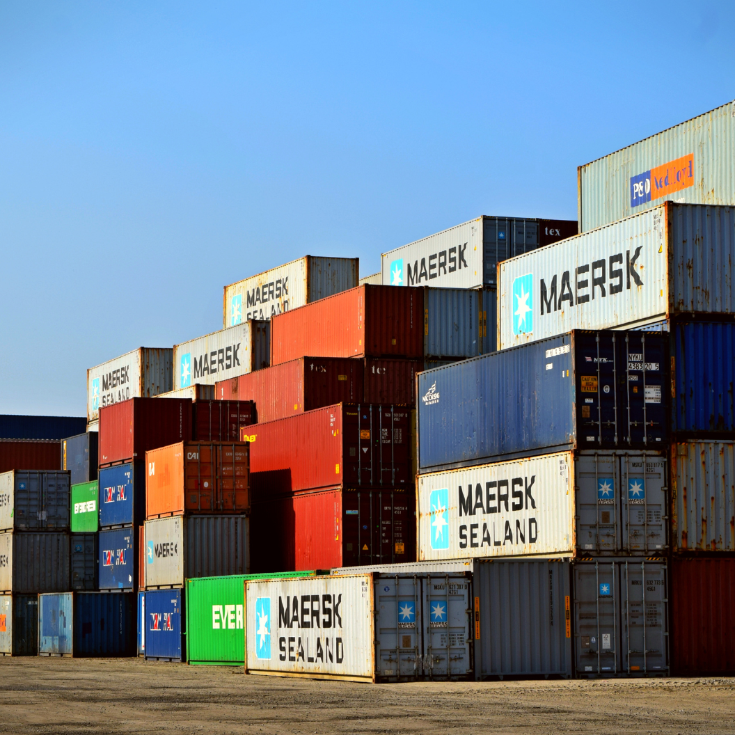 Stacked shipping containers in various colors including blue, red, green, and white, with prominent Maersk Sealand branding, set against a clear blue sky, illustrating global shipping and commodity market logistics.