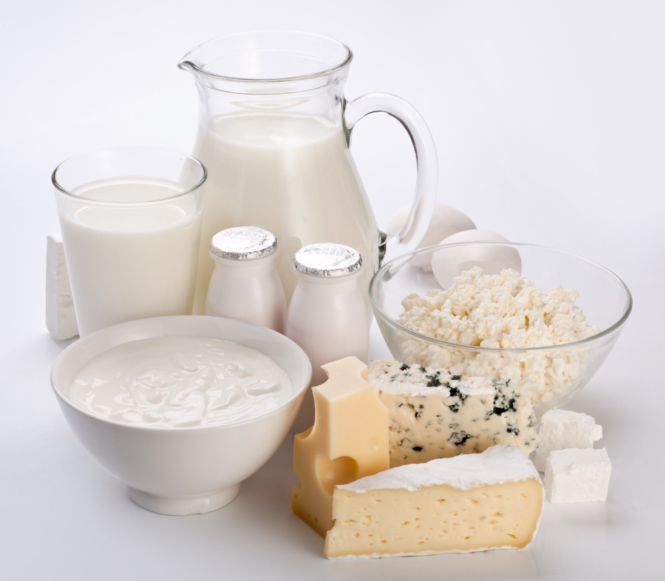 Assorted dairy products including a glass and pitcher of milk, yogurt in a bowl, two small sealed yogurt bottles, a bowl of cottage cheese, and various cheeses arranged on a white surface representing market insights and expert analysis for dairy commodities.