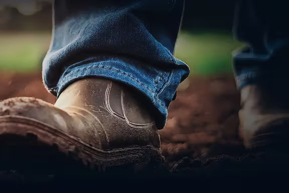 Close-up view of worn work boots stepping on dark soil outdoors, suggesting hands-on agricultural or fieldwork activity with a blurred natural background.