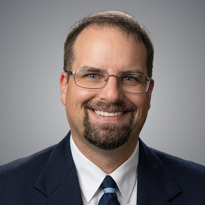 A man with short dark hair and a neatly trimmed beard wearing rectangular glasses, a dark suit, white shirt, and striped tie, smiling gently against a plain gray background.