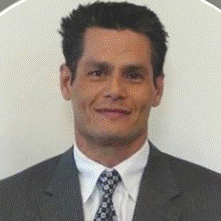 Man with dark hair wearing a gray suit and patterned tie, smiling with a neutral expression against a plain off-white background, conveying a professional and composed presence.
