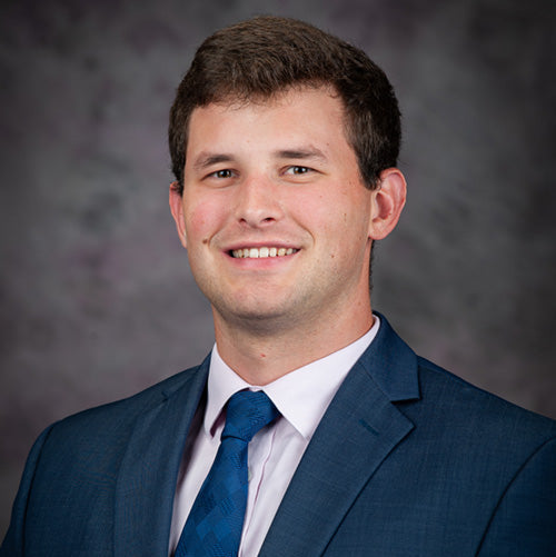 A professional headshot of a young man with short brown hair wearing a blue suit jacket, white dress shirt, and blue tie, set against a blurred dark gray background.