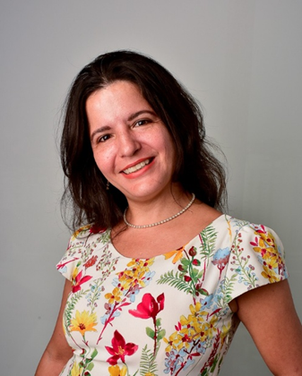 Woman with shoulder-length dark hair wearing a white blouse with colorful floral patterns, smiling warmly against a plain light gray background, conveying a friendly and approachable presence.