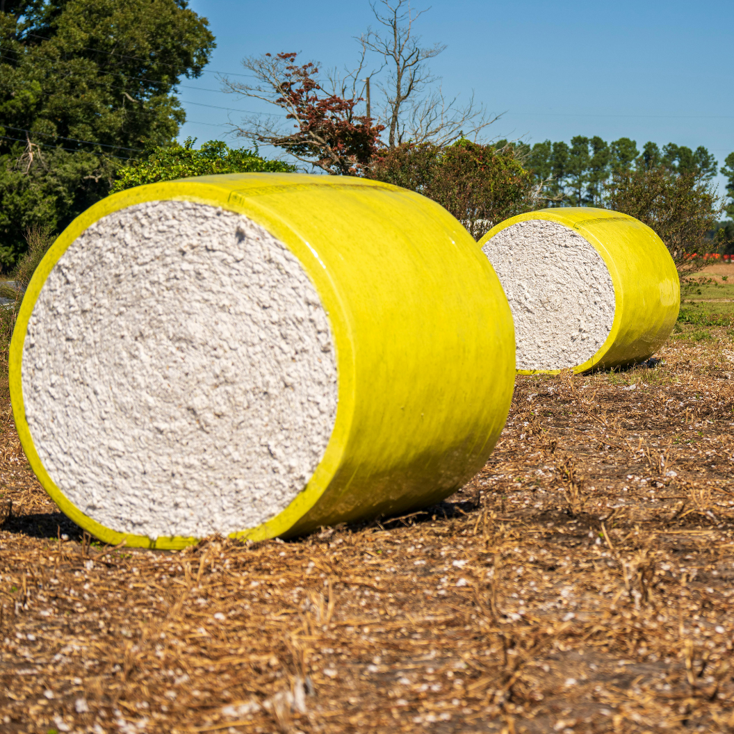 Two large cylindrical cotton bales wrapped in bright yellow plastic sit on a dry field with trees in the background, illustrating expert insights and exclusive market analysis for cotton commodity trends and Market Reports Subscriptions.