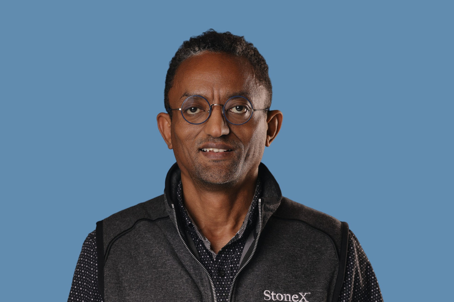 Portrait of a man wearing round glasses and a StoneX-branded vest over a dark shirt, set against a solid blue background for a professional market insights profile.