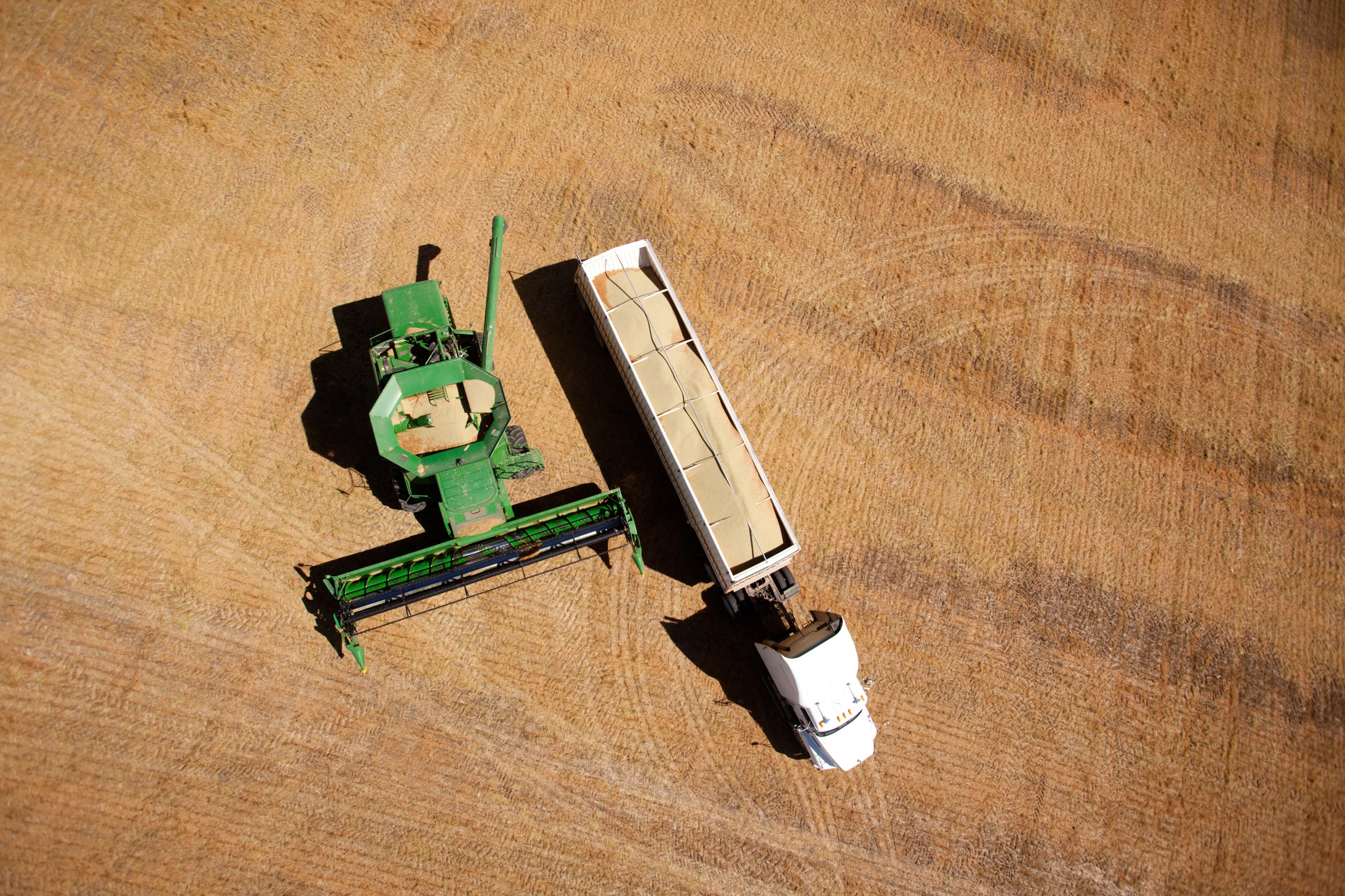 Aerial view of a green combine harvester unloading harvested grain into a white truck trailer on a vast, golden-brown field, illustrating commodity trends and market insights.