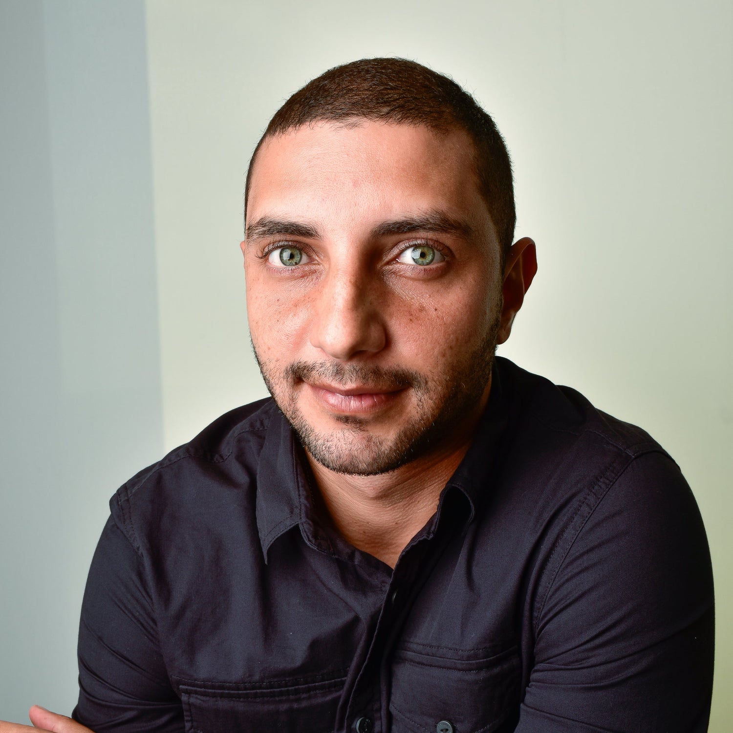Man with short dark hair and trimmed beard wearing a black button-up shirt, looking directly at the camera with a calm and confident expression against a neutral, softly lit background.