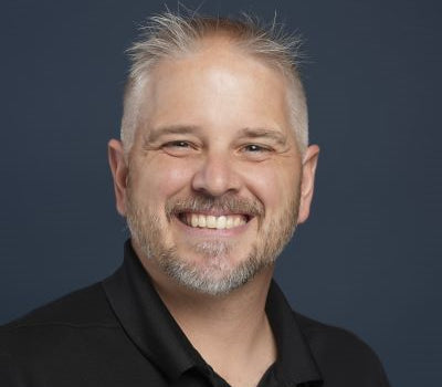 Close-up portrait of a smiling man with short gray hair and a trimmed beard, wearing a black shirt against a solid dark blue background, representing expert perspectives for Market Reports Subscriptions.