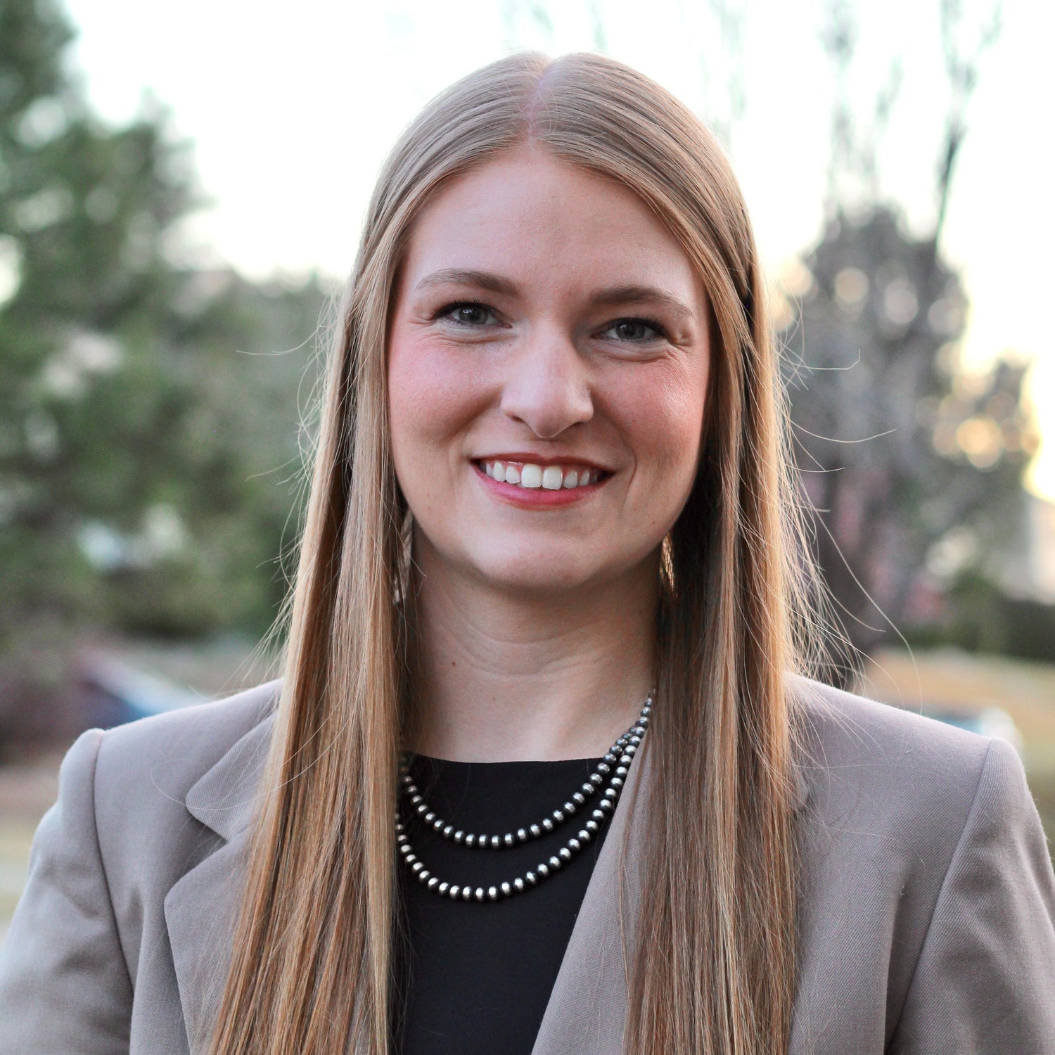 Woman with long straight blonde hair wearing a light gray blazer and layered black beaded necklace, smiling softly outdoors with blurred trees and soft sunlight in the background, conveying a professional and approachable presence.