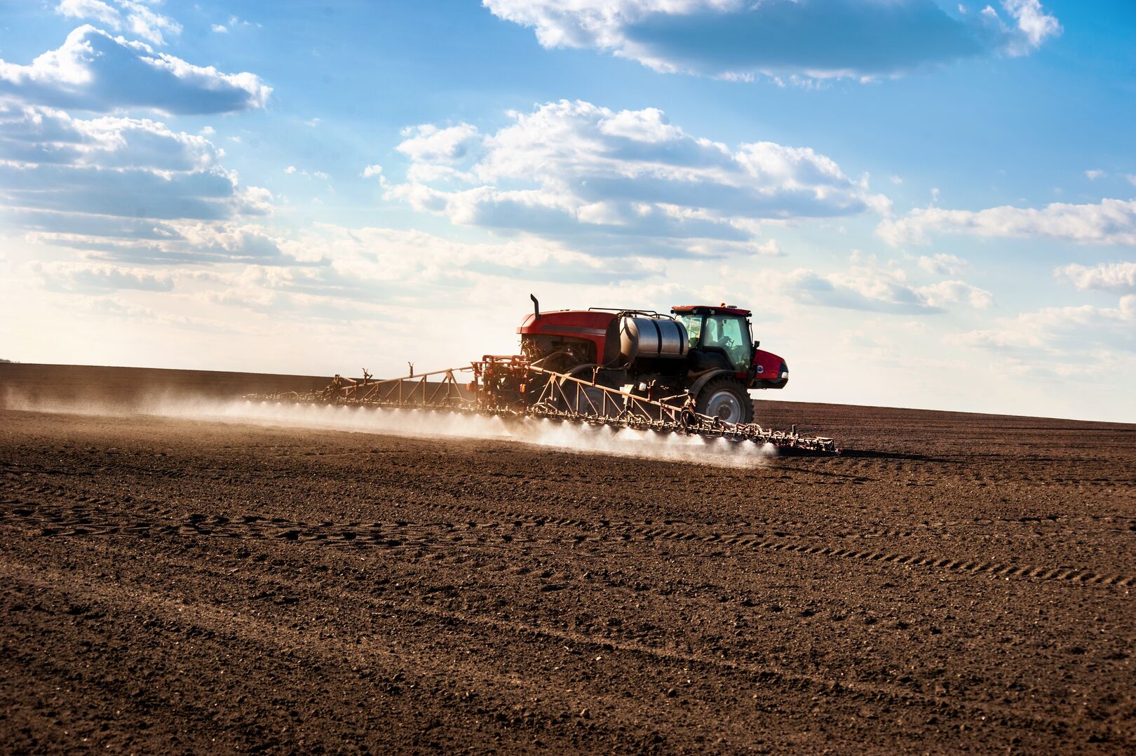 A red tractor equipped with a sprayer moves across a large, freshly tilled field under a partly cloudy sky, illustrating agricultural commodity production and farming activity.