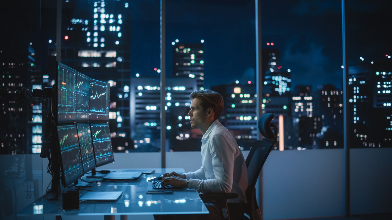 A focused man in a white shirt analyzing multiple financial market charts on large computer screens in a modern office with a nighttime city skyline visible through the windows.