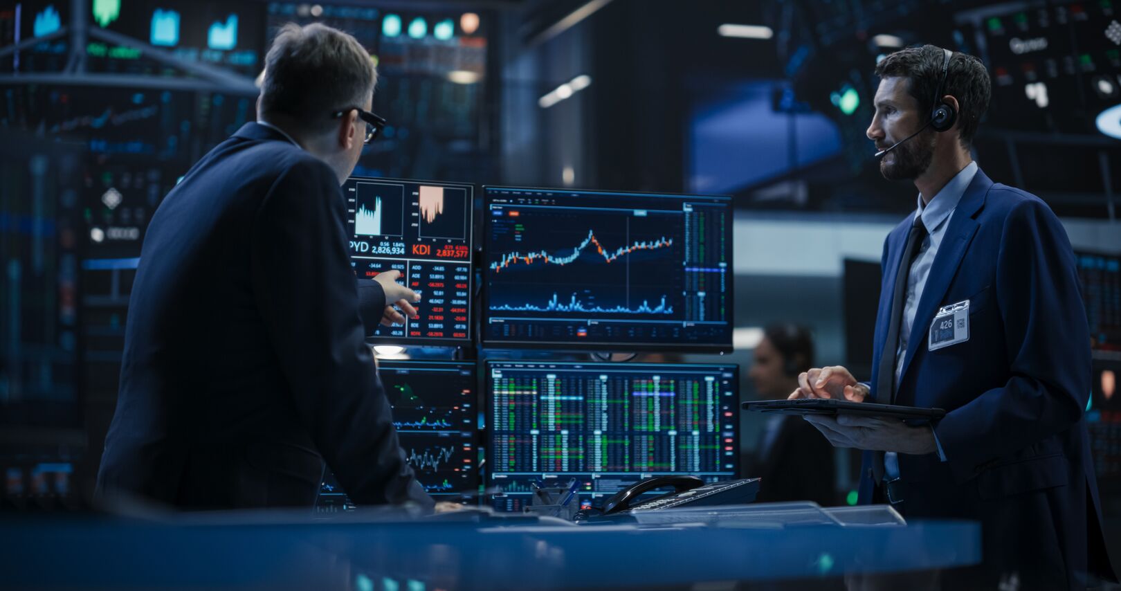 Two professionals in a dimly lit trading room review multiple monitors displaying financial charts and market data, discussing market trends and analysis.