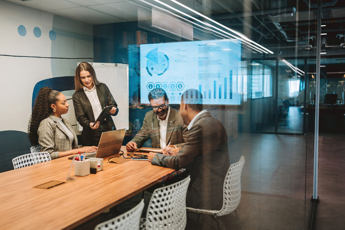 A diverse group of four business professionals collaborating around a conference table with a laptop and documents, with charts and graphs visible on a glass wall in a modern office setting.