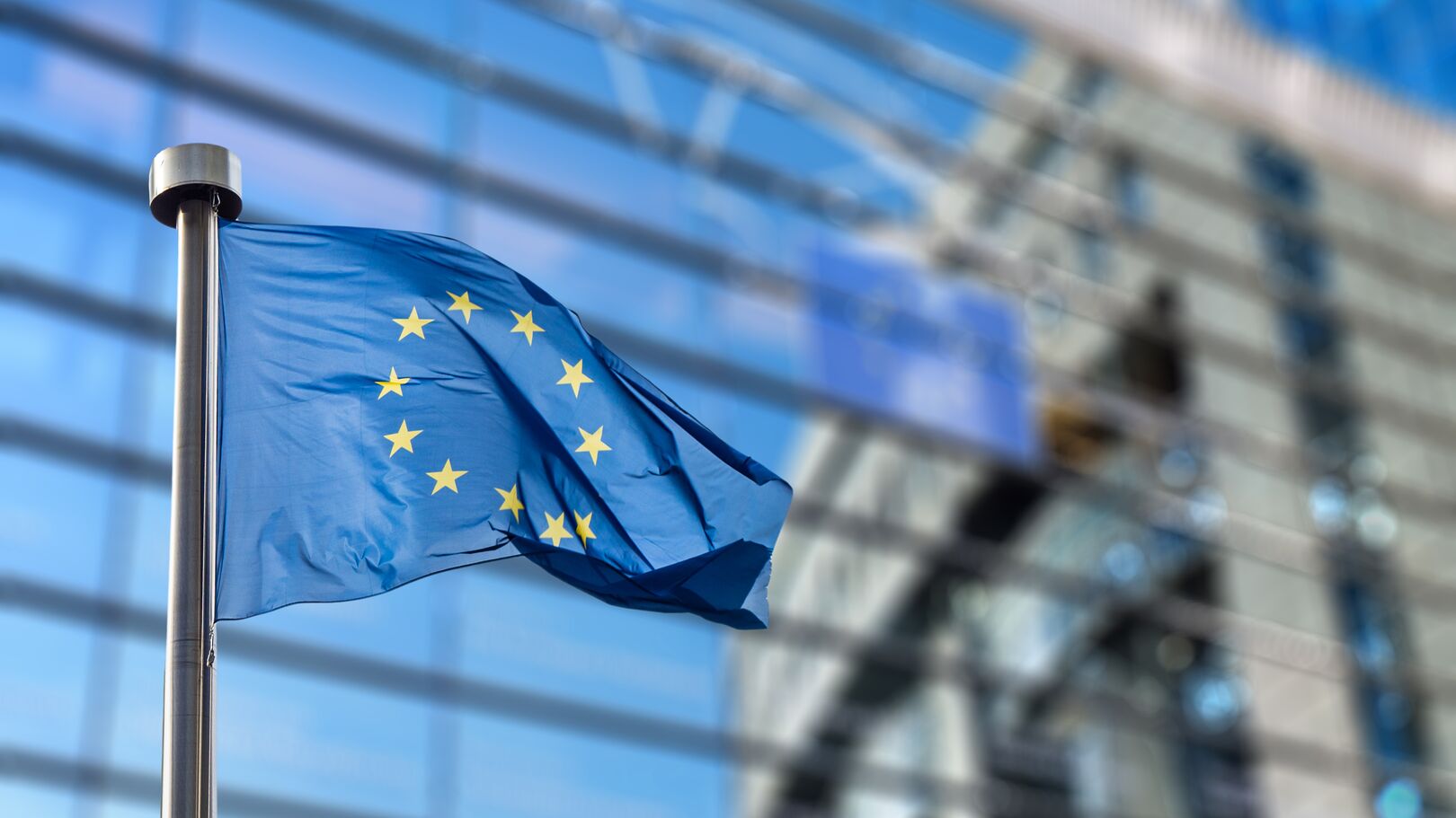 European Union flag fluttering on a flagpole in front of a modern glass building with reflections, symbolizing international market dynamics and economic perspectives.