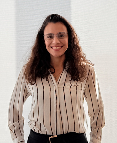 Woman with long curly dark hair and glasses wearing a white blouse with black vertical stripes, smiling confidently against a bright softly lit background, conveying a professional and approachable presence.