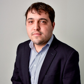 Man with short dark hair and light stubble, wearing a dark blazer and light blue shirt, posing with a neutral expression against a plain light gray background, conveying a calm and professional presence.