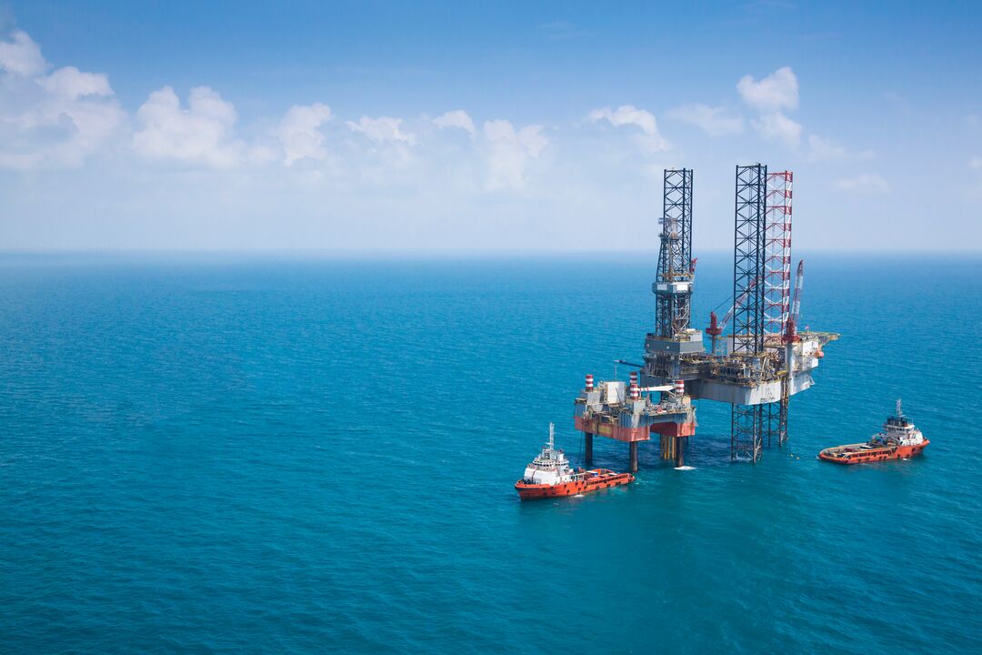 An offshore oil drilling platform surrounded by blue ocean water with two supply vessels positioned nearby under a partly cloudy sky.