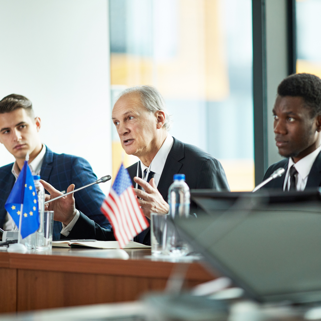 Three men in business attire participate in a formal discussion around a conference table with European Union and United States flags visible, highlighting global macroeconomic perspectives and expert insights for market reports subscriptions.