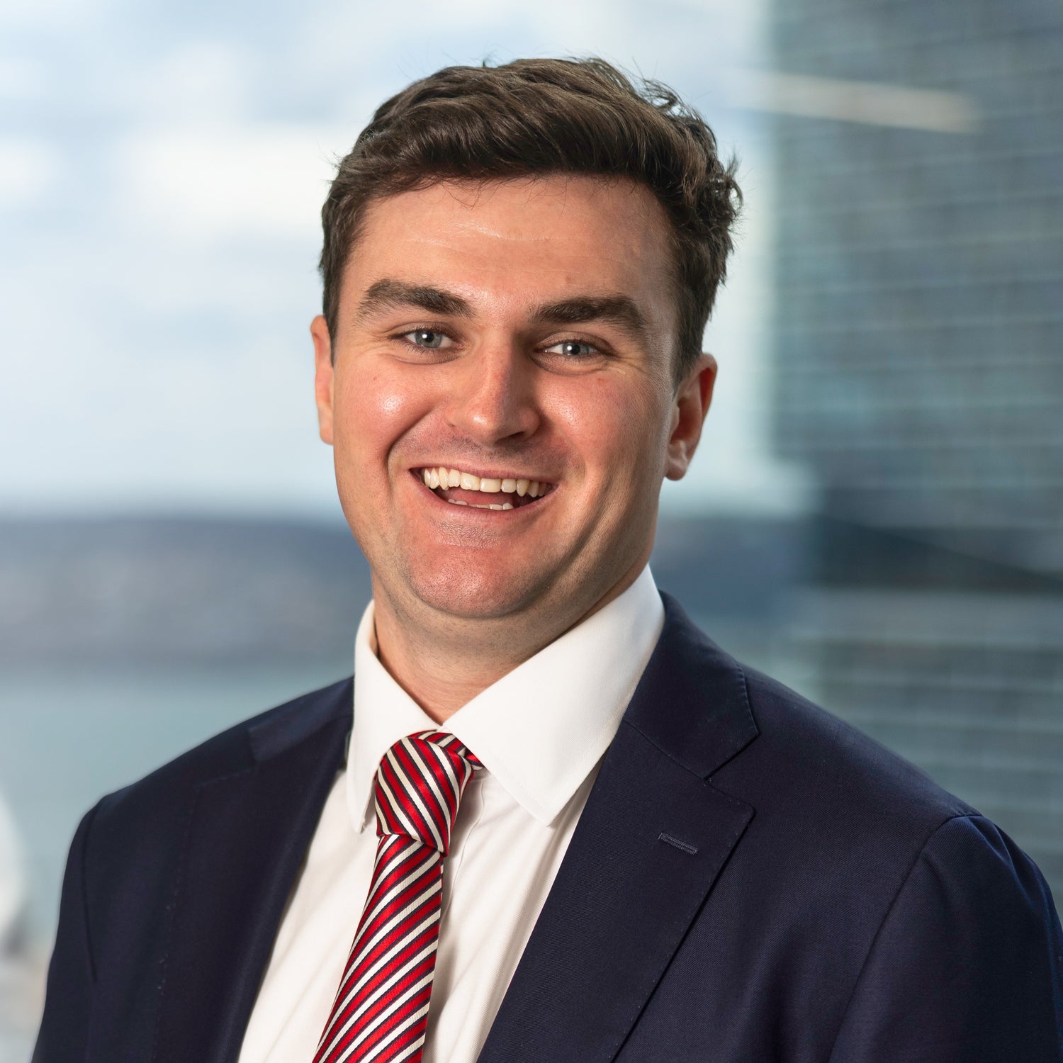 A young man with short brown hair wearing a dark suit jacket, white shirt, and red-striped tie, smiling warmly with a blurred cityscape and body of water in the background.