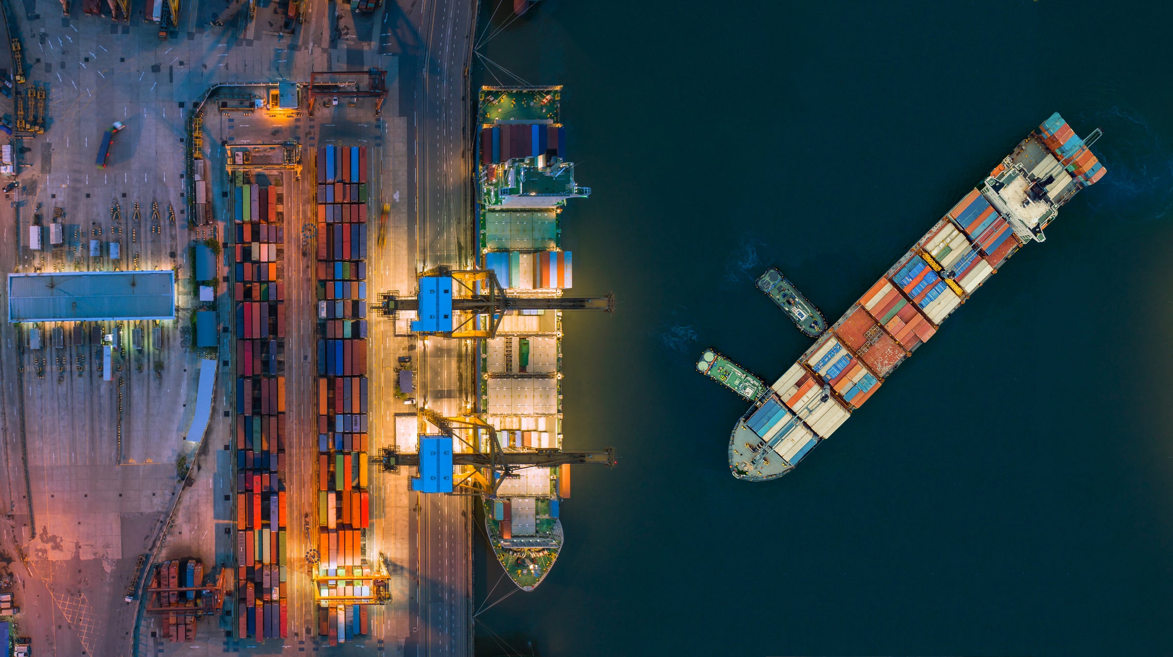Aerial nighttime view of a busy port with two container ships docked and brightly lit cranes loading and unloading colorful shipping containers, illustrating global commodity flow and market trends.