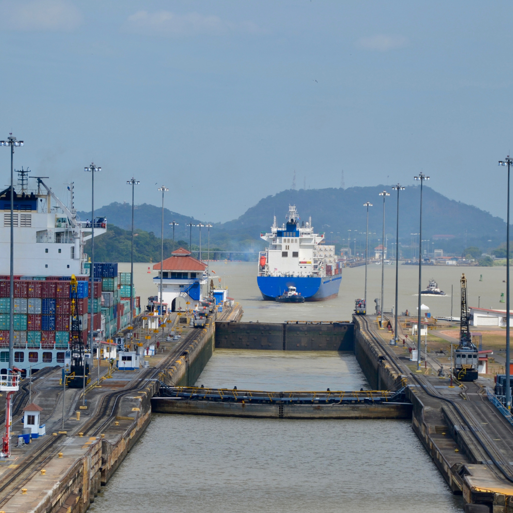 View of a large cargo ship passing through a canal lock, surrounded by container stacks and port infrastructure with hills visible in the distance, symbolizing global trade flow and market trends for Market Reports Subscriptions with expert insights and exclusive market analysis.