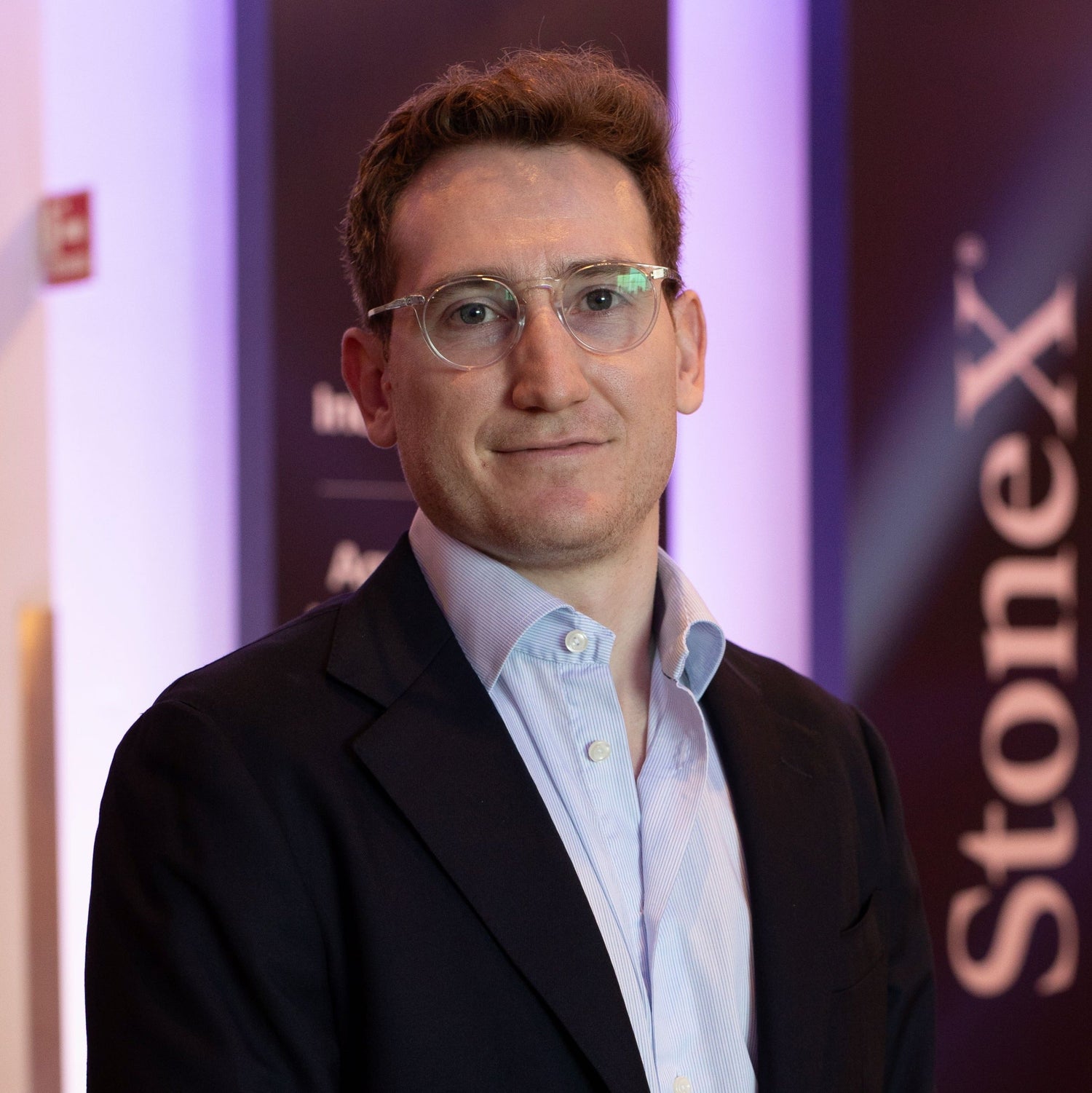 Man with short curly hair and clear glasses wearing a light blue shirt and dark blazer, posing with a neutral expression in front of a backdrop featuring the StoneX logo and market insights branding.